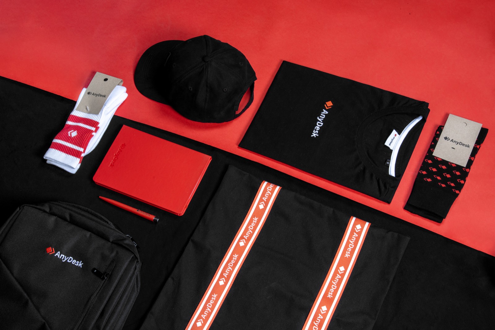 Een flatlay van zwarte en rode merchandise: een pet, een opgevouwen T-shirt, een rood notitieboek met pen, twee paar sokken, een rugzak en een zwart item met gestreepte rand.