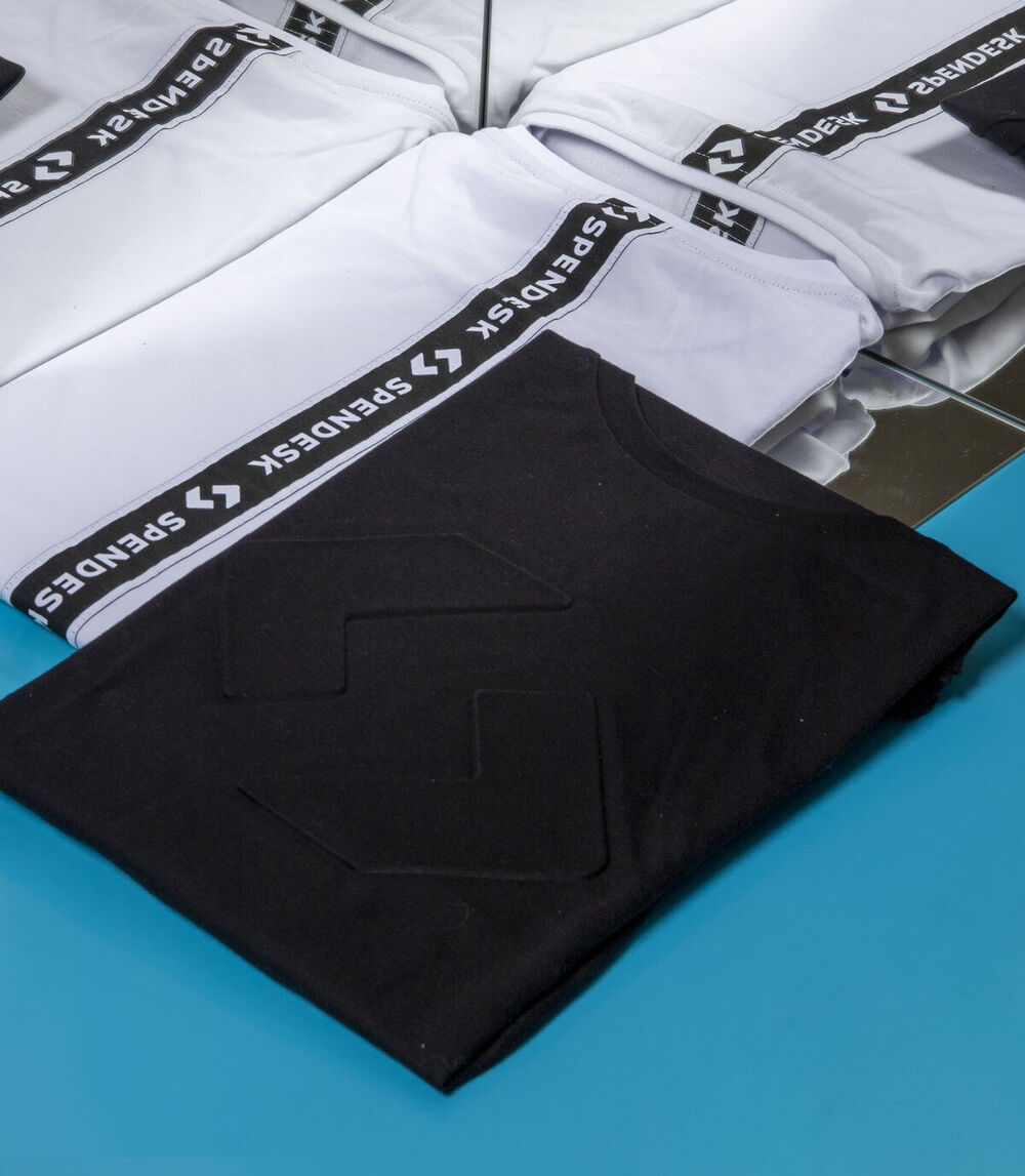 Folded black T-shirt with embossed logo on top of white garments with black Spendesk logo tape along the sleeves, on a blue surface