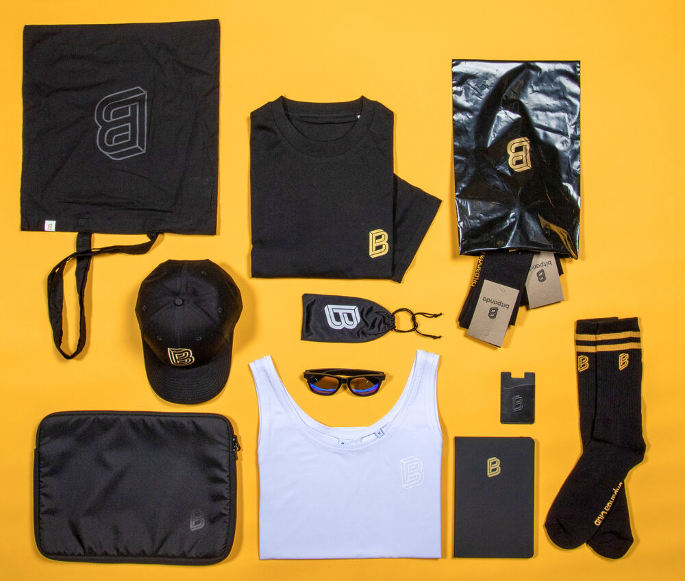 A flat-lay of the Bitpanda merchandise collection: tote, t-shirt, cap, socks, laptop sleeve, notebook, sunglasses and card holder in black and gold on a yellow background