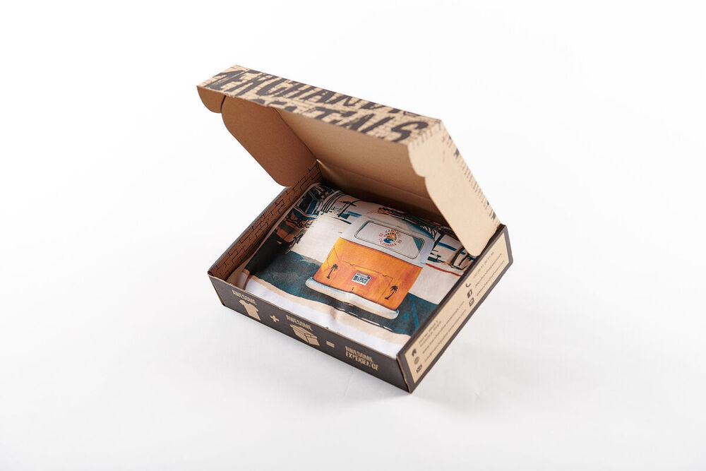 Open printed mailer box with a 'Merchandise Rentals' lid, holding a folded white T-shirt printed with an orange camper van