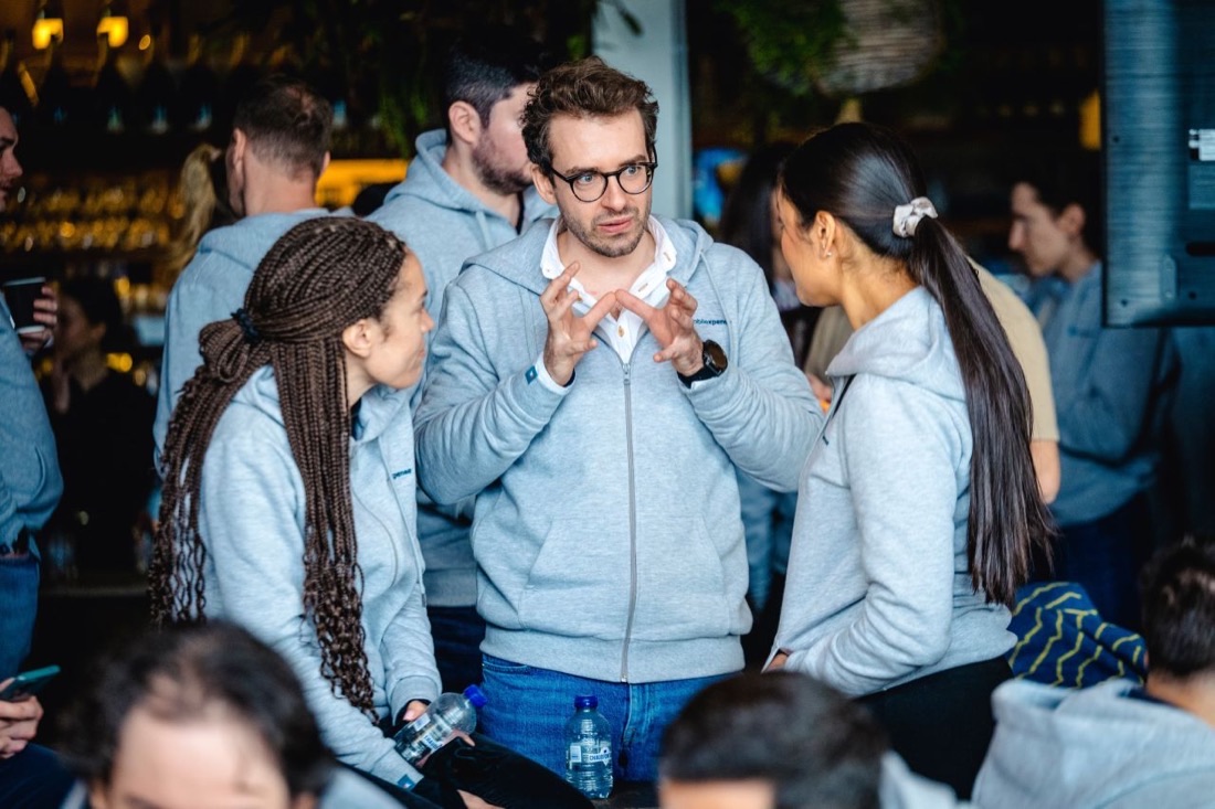 Collega's in bijpassende lichtgrijze zip-hoodies met een klein logo op de borst praten op een drukke, schemerig verlichte locatie, de man in het midden gebaart terwijl hij praat.
