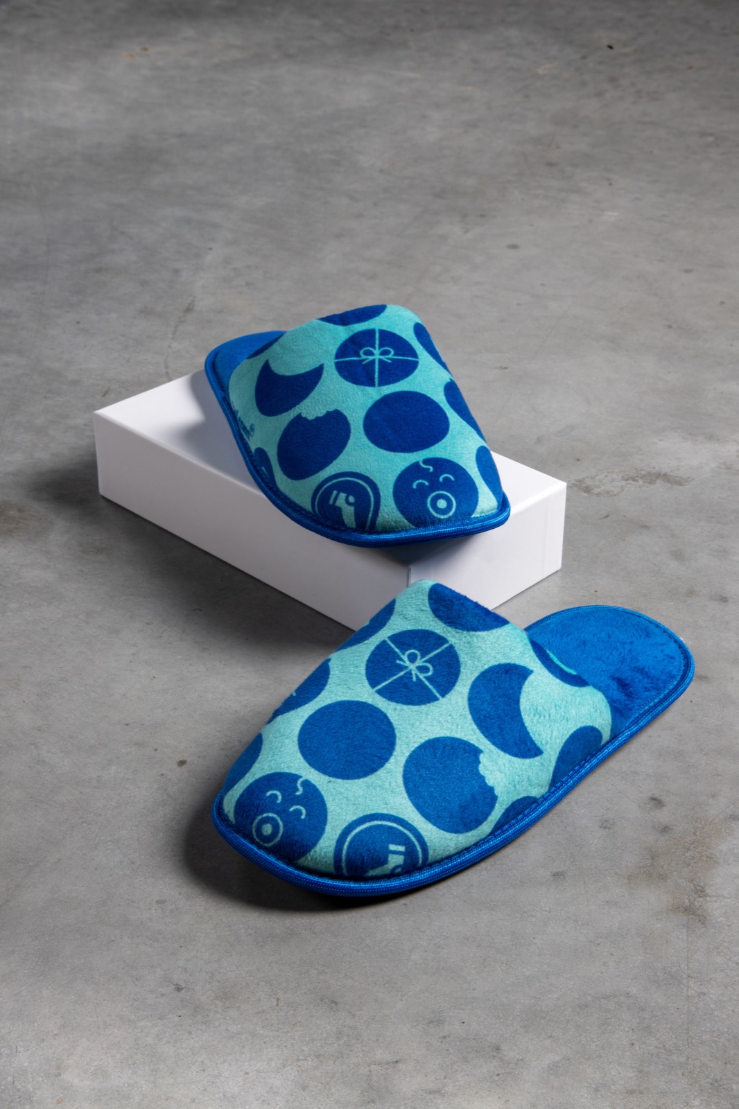 Een paar blauw-turquoise pantoffels met een groot stippenpatroon, een ervan rustend op een witte doos, op een betonnen vloer