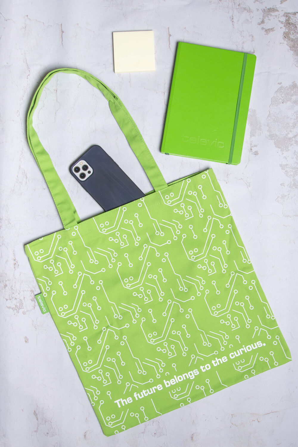 A green branded tote bag with a circuit-pattern print and brand slogan, laid out with a matching branded notebook and a phone
