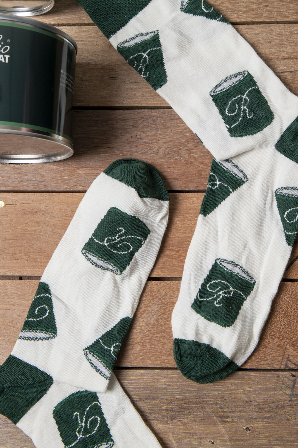 Cream branded socks with a repeating green product-tin motif, photographed next to the manufacturer's actual product tin on a wooden surface