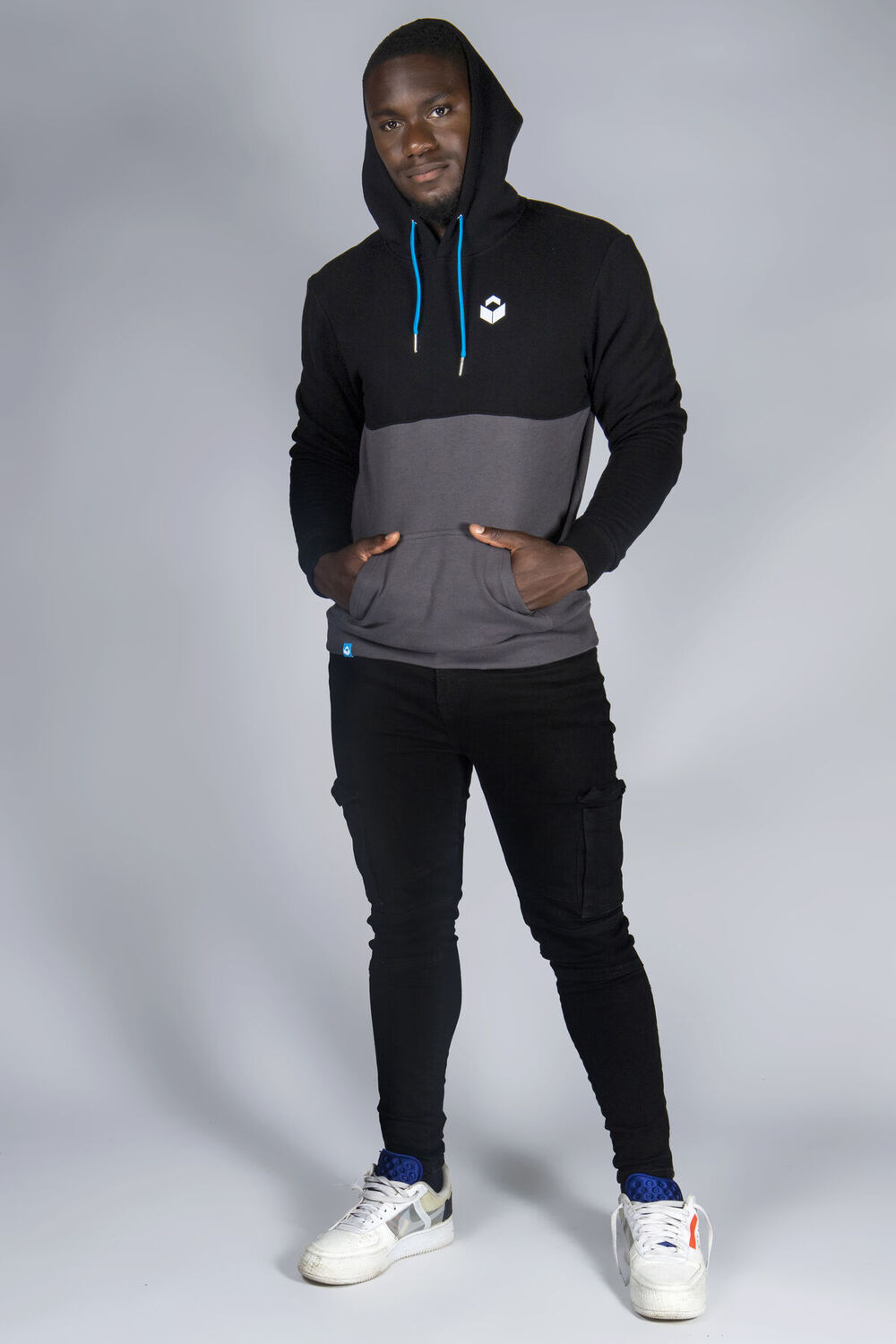 Man standing in a studio with hands in the pocket of a black and grey colour-block hoodie with blue drawcords and a small white chest logo