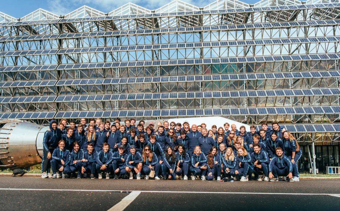 Een team van meer dan vijftig mensen in bijpassende marineblauwe trainingspakken poseert in rijen voor een grote glazen kas vol zonnepanelen.