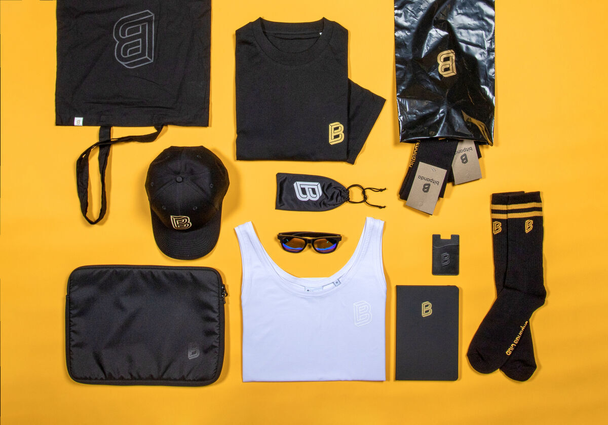 Bitpanda merchandise flat lay on yellow: black tote, T-shirt and cap with gold B logo, white tank top, sunglasses, notebook, laptop sleeve and socks