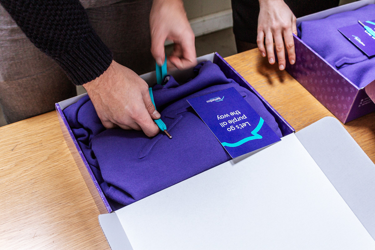 Het uitpakken van een paarse gebrande kerstcadeaudoos, met een paarse trui en een kaartje met de tekst 'Let's go purple all the way'