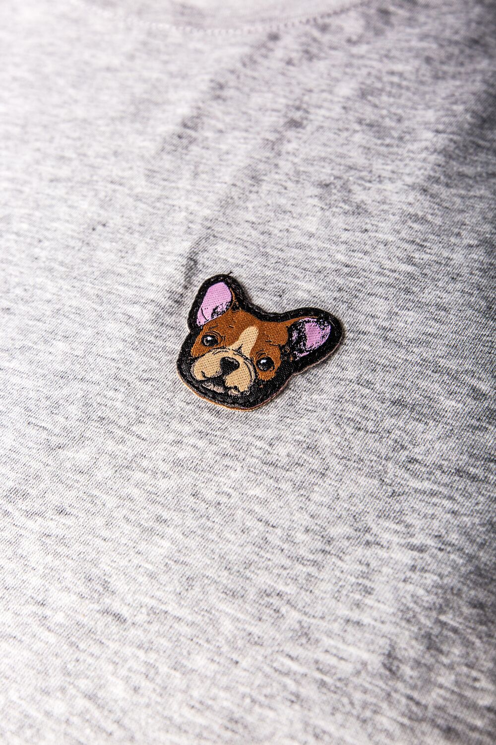 Una camiseta jaspeada gris con un pequeño parche tejido de personaje, ejemplo de una pieza de colección distintiva que puede encajar mejor en una campaña que en una tienda del día a día