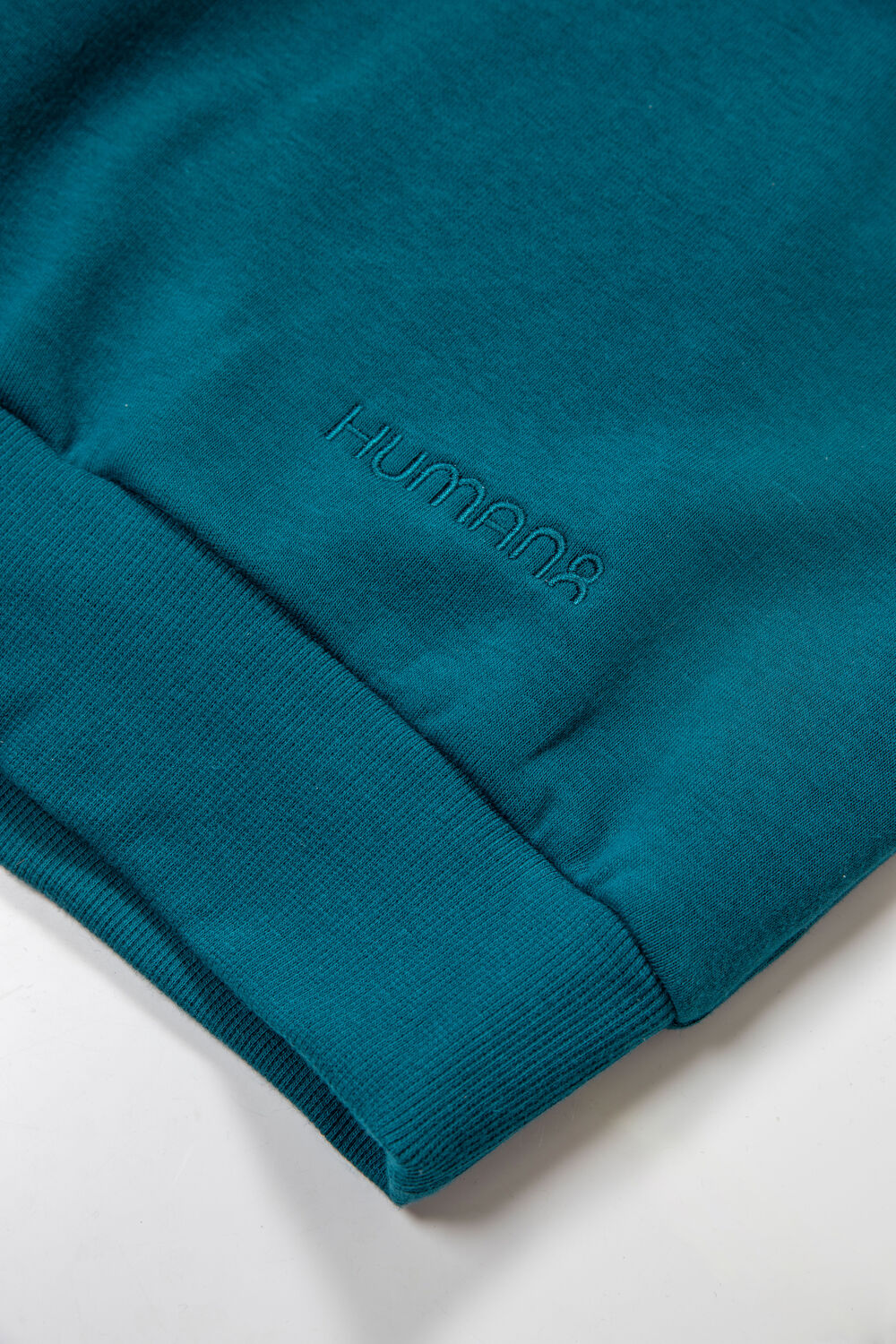 Primer plano del puño y el bajo de una sudadera color teal con un logo bordado tono sobre tono discreto, un producto mantenido como stock de almacén