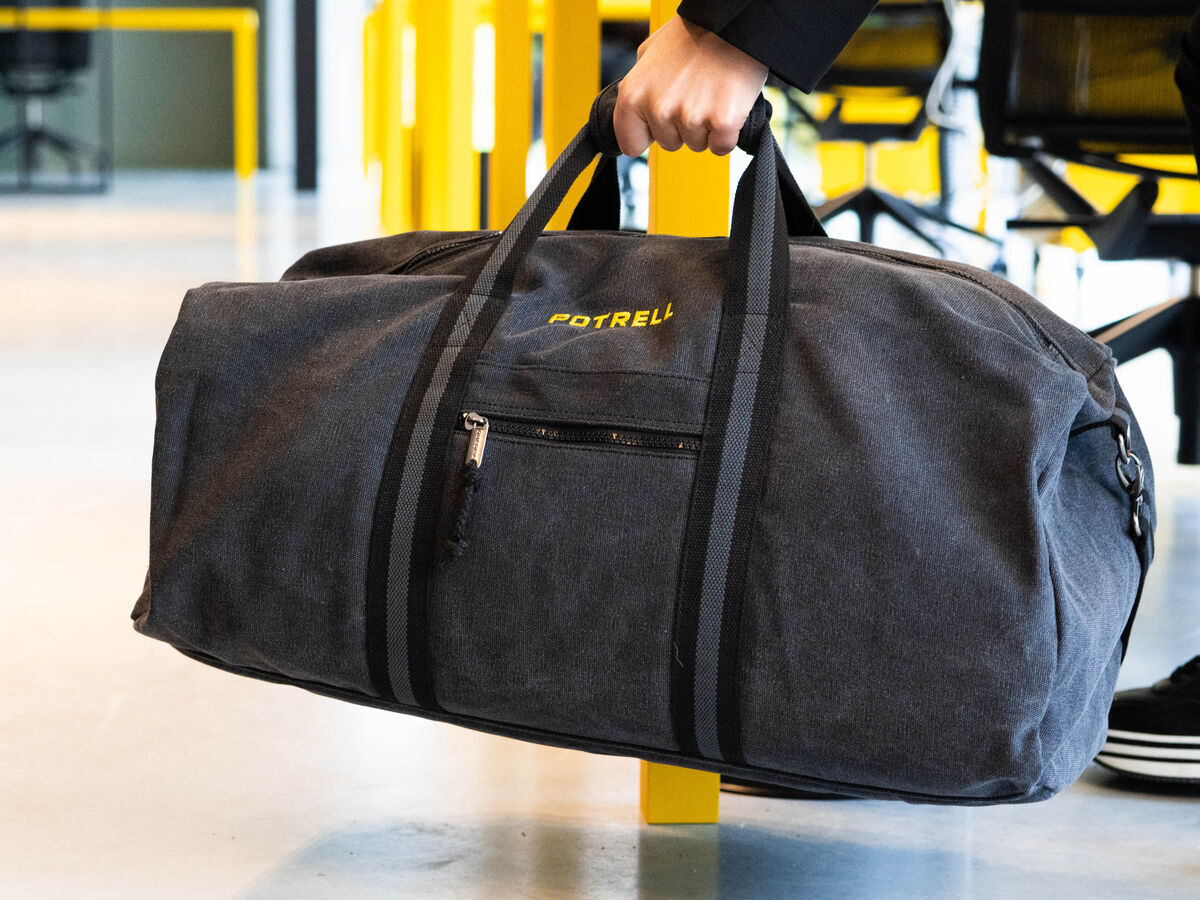 A subtly branded charcoal canvas weekend bag being carried through an office, the kind of corporate gift that keeps generating impressions long after a food gift would have been eaten
