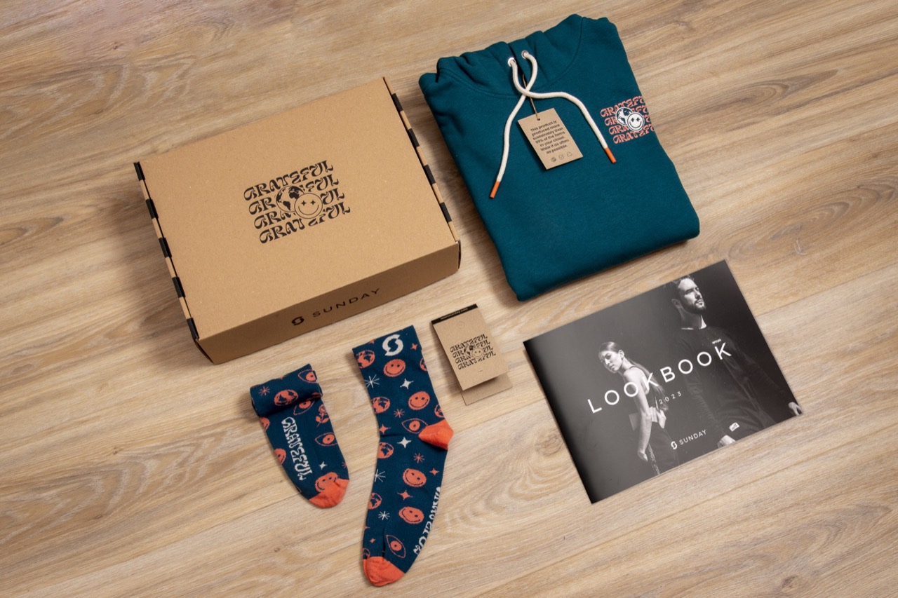 Een kartonnen geschenkdoos, een opgevouwen petrolkleurige hoodie, een paar sokken met patroon, een hangtag en een gedrukt lookbook uitgelegd op een houten vloer