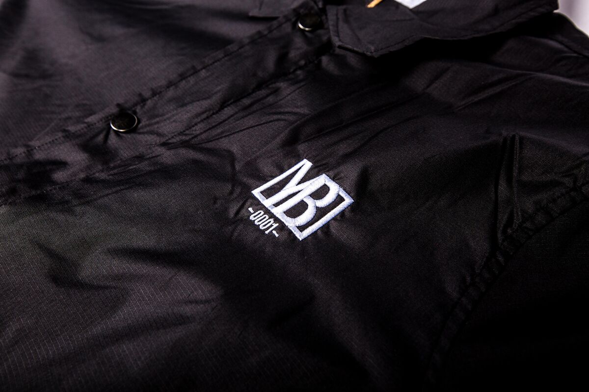Detalle de una chaqueta ripstop negra con un pequeño logo bordado en blanco y botones de presión, parte de una colección de merchandising producida y conservada como stock de almacén
