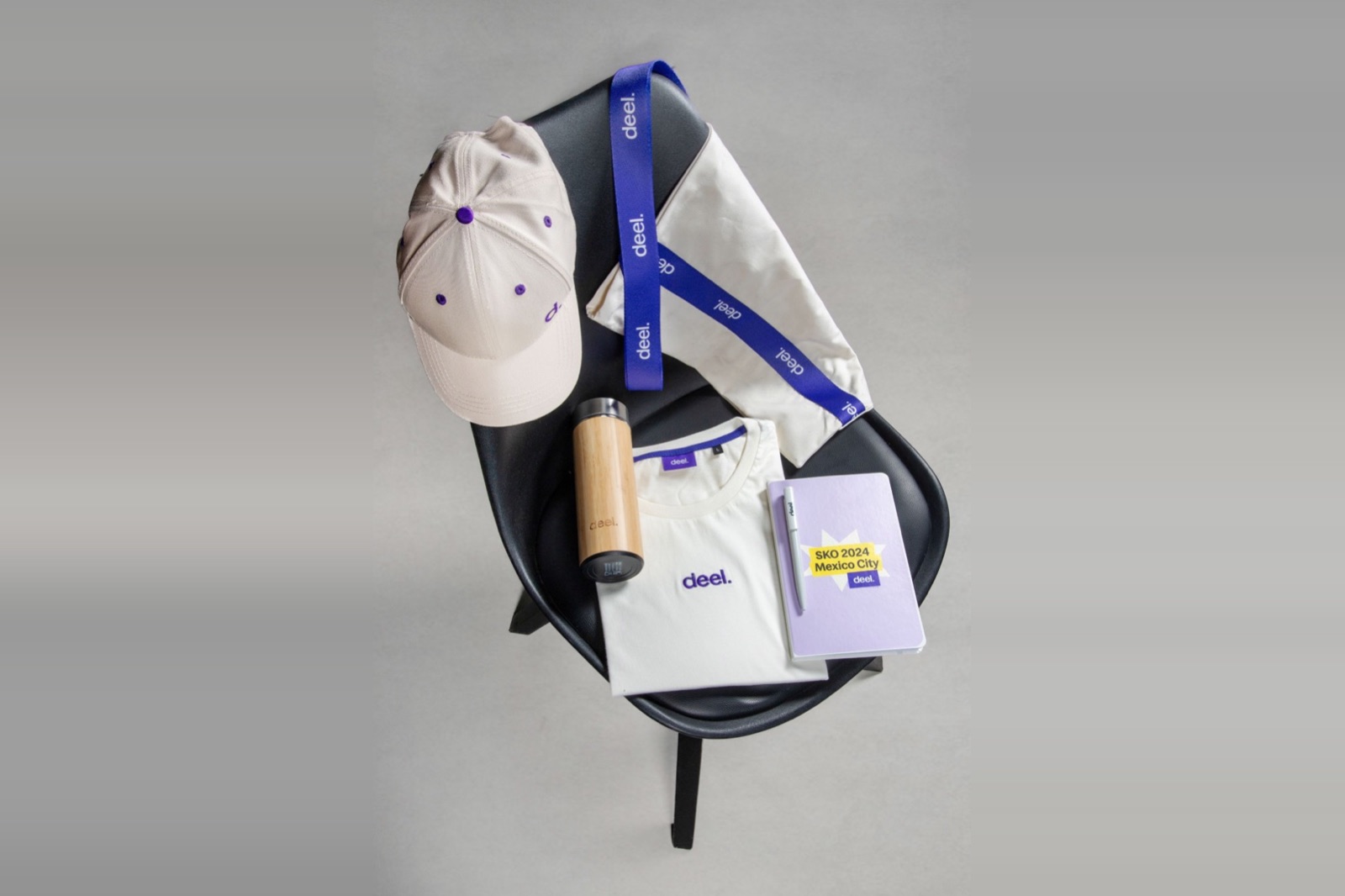 Een SKO-pack uitgestald op een zwarte stoel: een crèmekleurige pet, een blauwe lanyard, een tote bag, een bamboefles, een opgevouwen T-shirt en een lila notitieboek met pen.