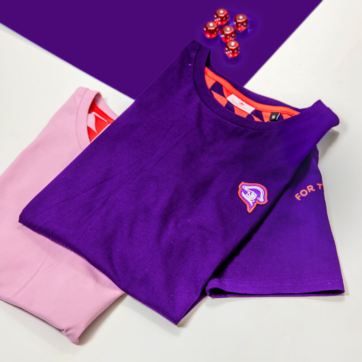 Dos camisetas de marca dobladas de una misma colección aprobada, una con un parche bordado en el pecho y otra con una manga estampada, producidas según la misma especificación de marca