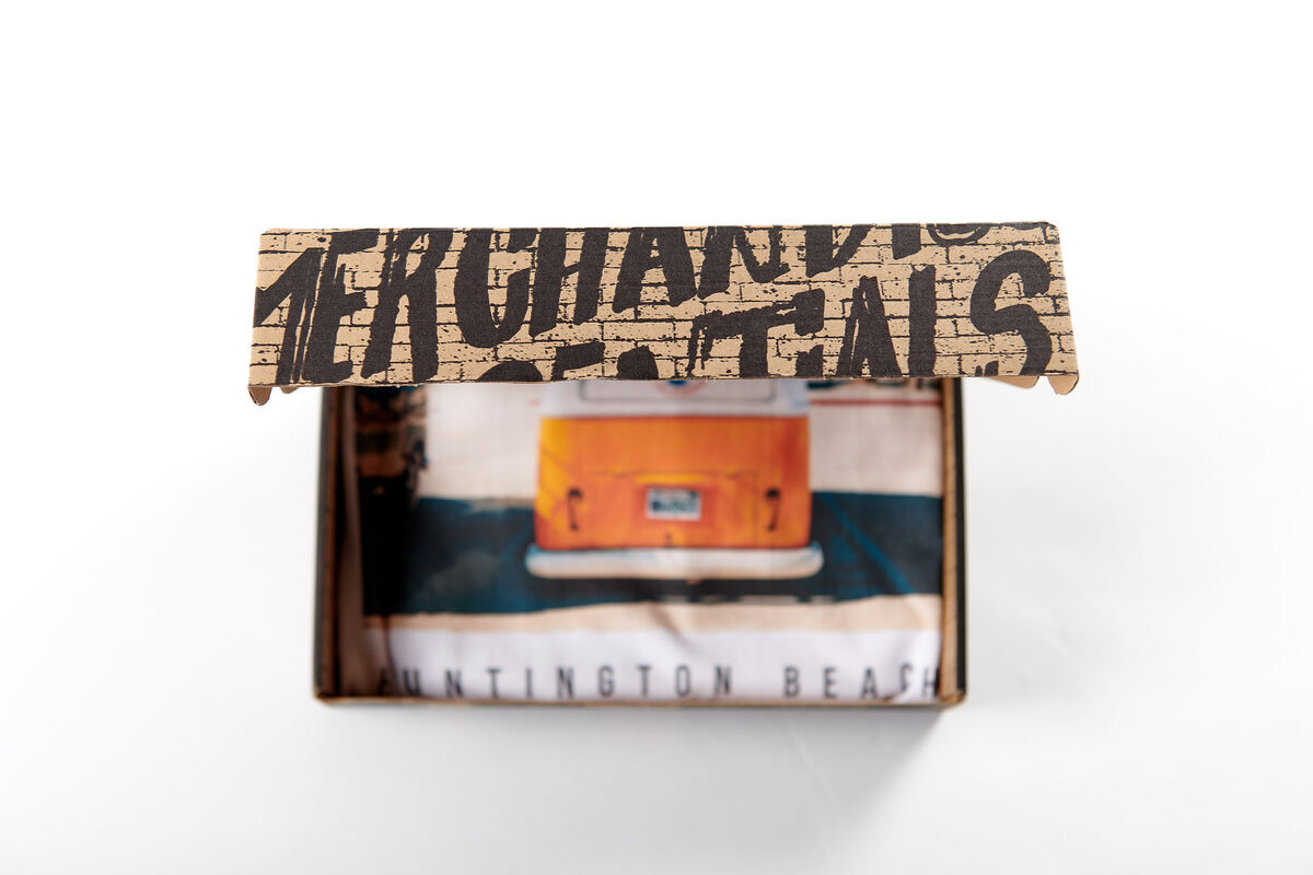 Front view of an open mailer box with a printed 'Merchandise Rentals' lid, showing a folded T-shirt with an orange camper van and 'Huntington Beach' text